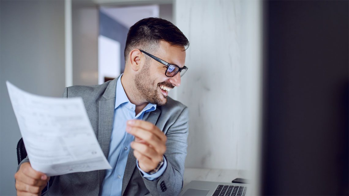 Man smiling at a computer screen holding a piece of paper wearing a gray suit and glasses.