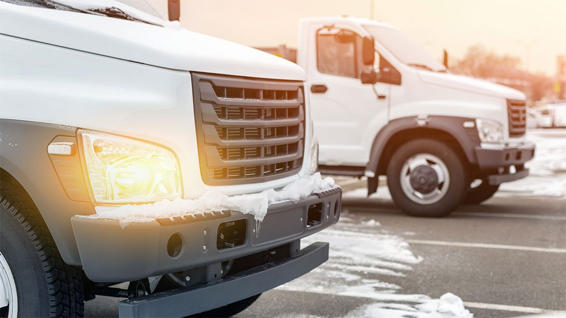 Two white tuck fleet vehicles covered in snow running with their headlights on.