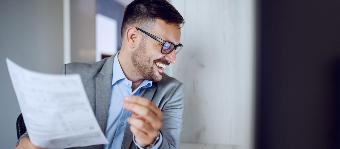 Man smiling at a computer screen holding a piece of paper wearing a gray suit and glasses.