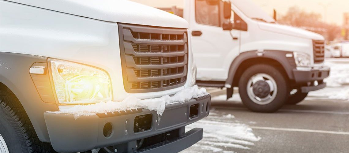 Two white tuck fleet vehicles covered in snow running with their headlights on.